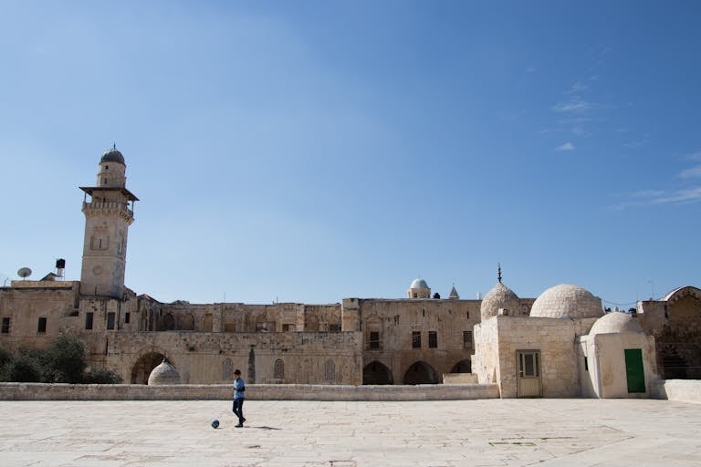 A young boy kicking a ball in front of an ancient mosque with domes and towers under a clear blue sky.