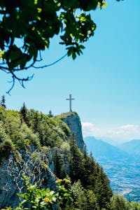 A scenic mountain landscape with a prominent cross, surrounded by dense green foliage under a clear blue sky.