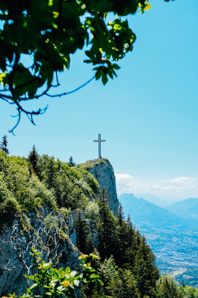 A scenic mountain landscape with a prominent cross, surrounded by dense green foliage under a clear blue sky.